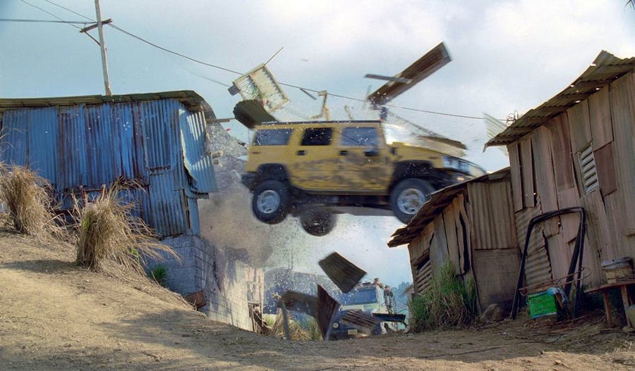 A bright yellow Humvee flies through the air between two shacks made of corrugated metal sheets.