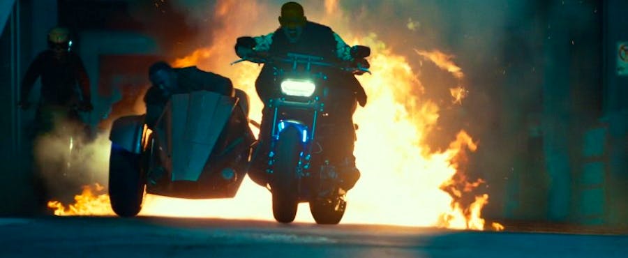 Mike rides a motorcycle, screaming, while Marcus cowers in the sidecar. Behind them, something explodes.