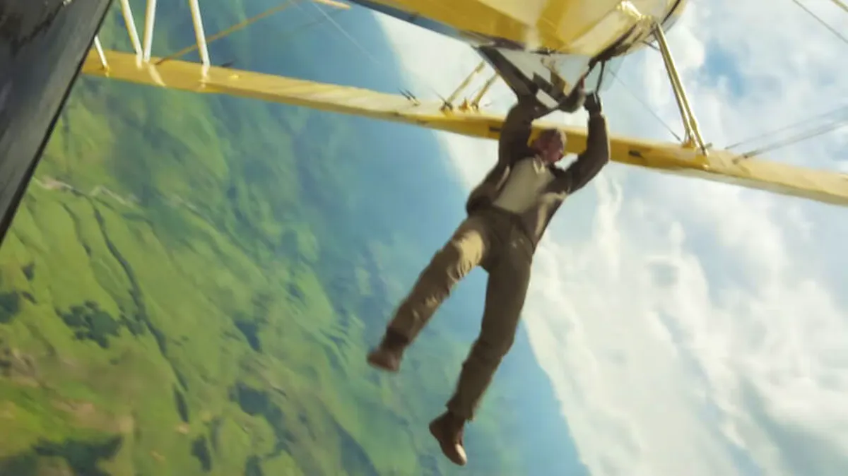 A man hangs precariously from the struts of a biplane