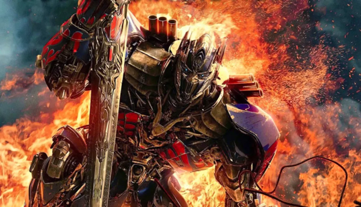 Optimus Prime kneels with a sword as something explodes behind him